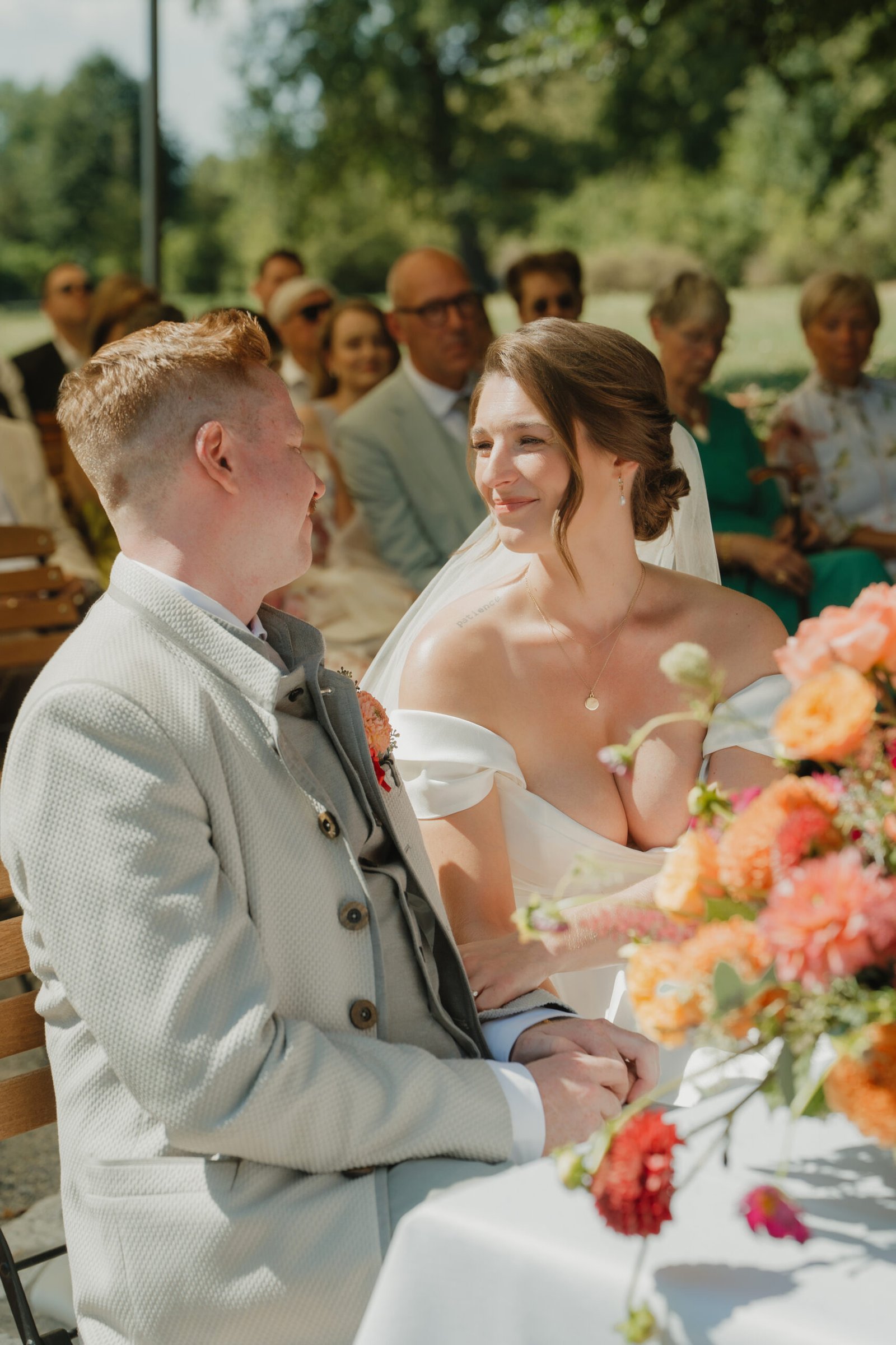 Braut und Bräutigam bei der freien Trauung mit Blumenstrauß und Gästen im Hintergrund